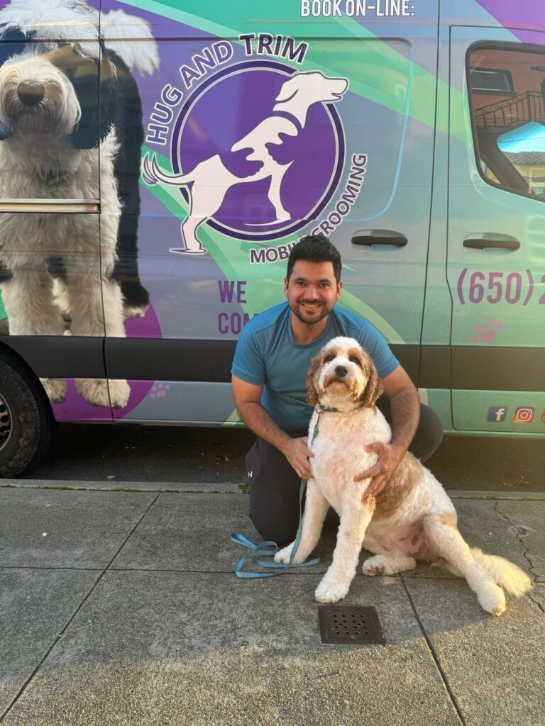 Man kneeling beside a large dog on a sidewalk in front of a van featuring a dog grooming service advertisement.
