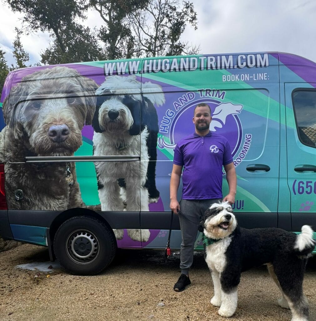 A man in a purple shirt stands beside a black and white dog in front of a van with pet grooming advertisements featuring images of dogs.